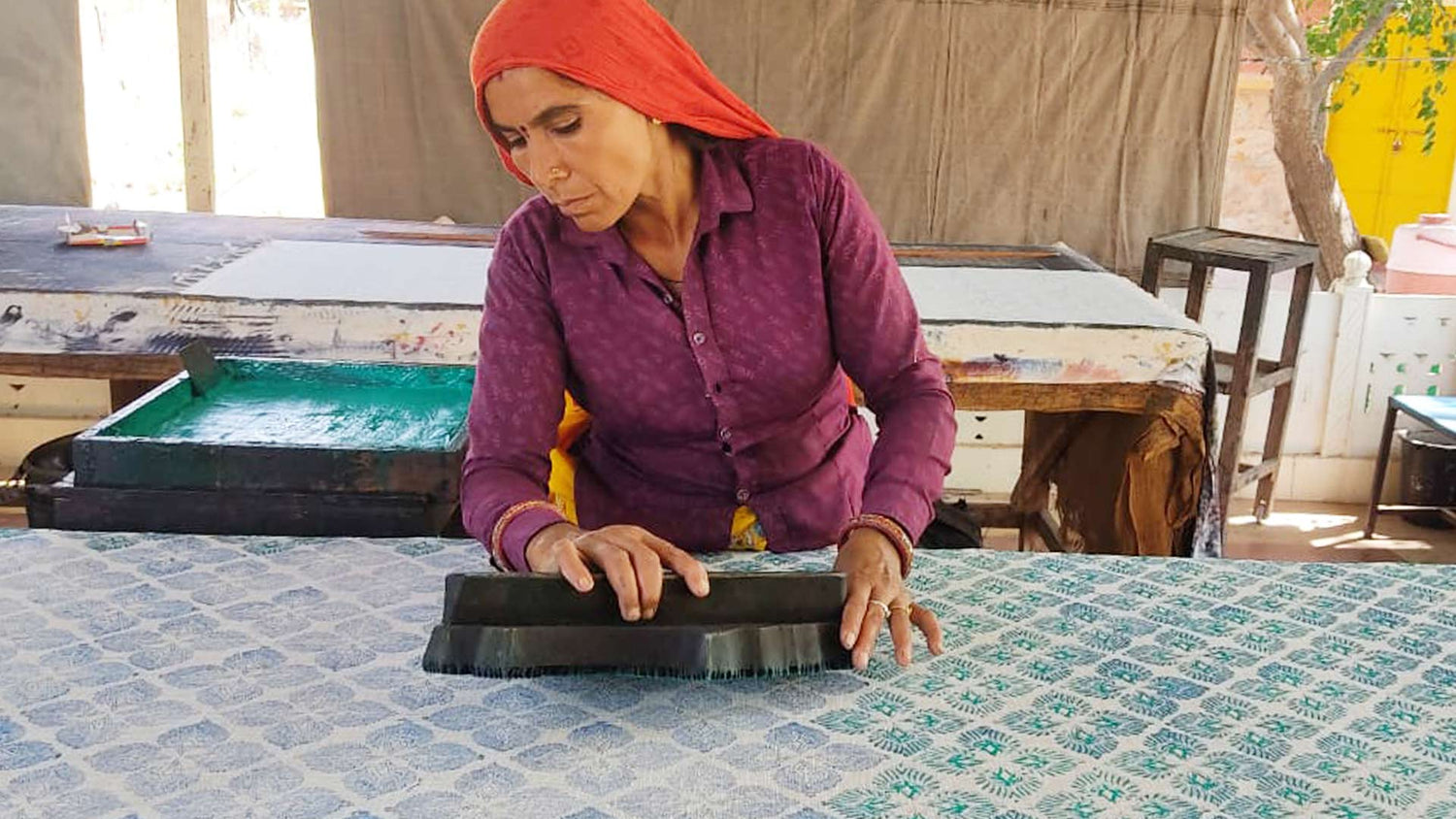 Indian woman in a bright orange headscarf using a big wooden block to print fabric.