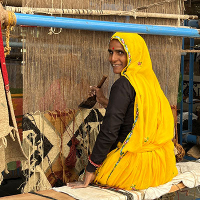 artisan wearing a bright yellow sari and weaving a rug on a vertical loom.