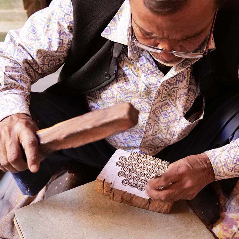 Artisan carving a floral design onto a wooden block with a small chisel and a wooden stick.