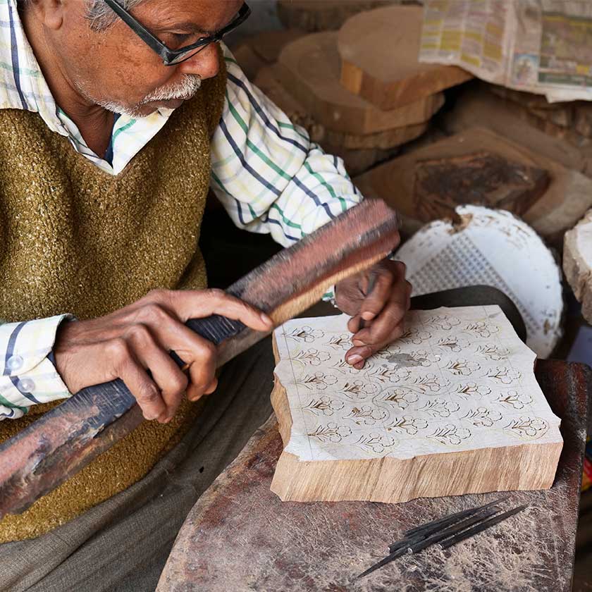 Man using a small chisel and a big wooden plank to carve a design onto a block of wood.