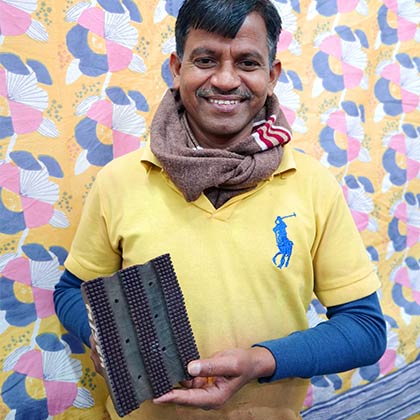 Artisan holding a wooden block for printing, in front of a floral pattern sheet.