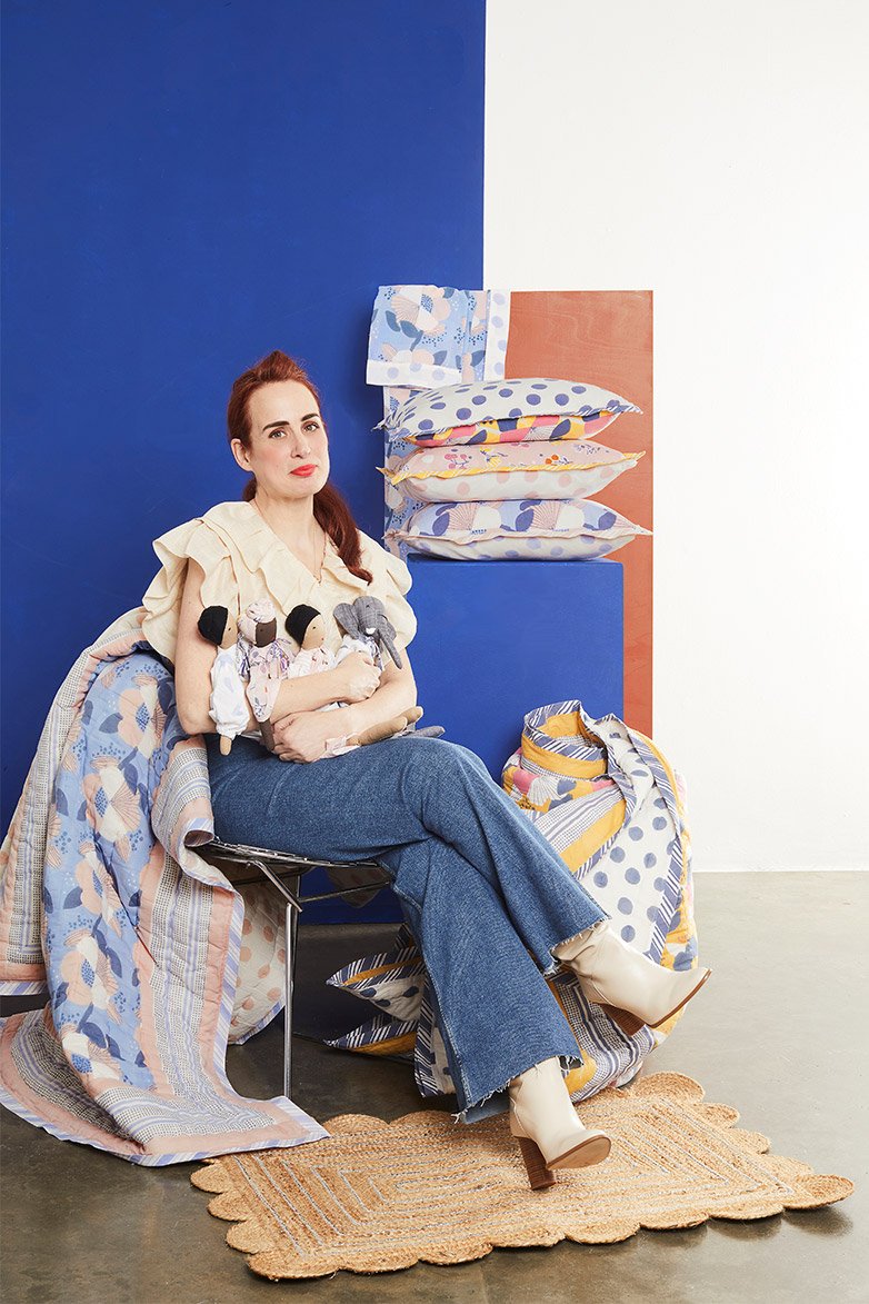 Founder Noa Alvarez sittin on a chair and surrounded by products from the collection including printed quilts, cushions, four dolls and a rug in the floor.