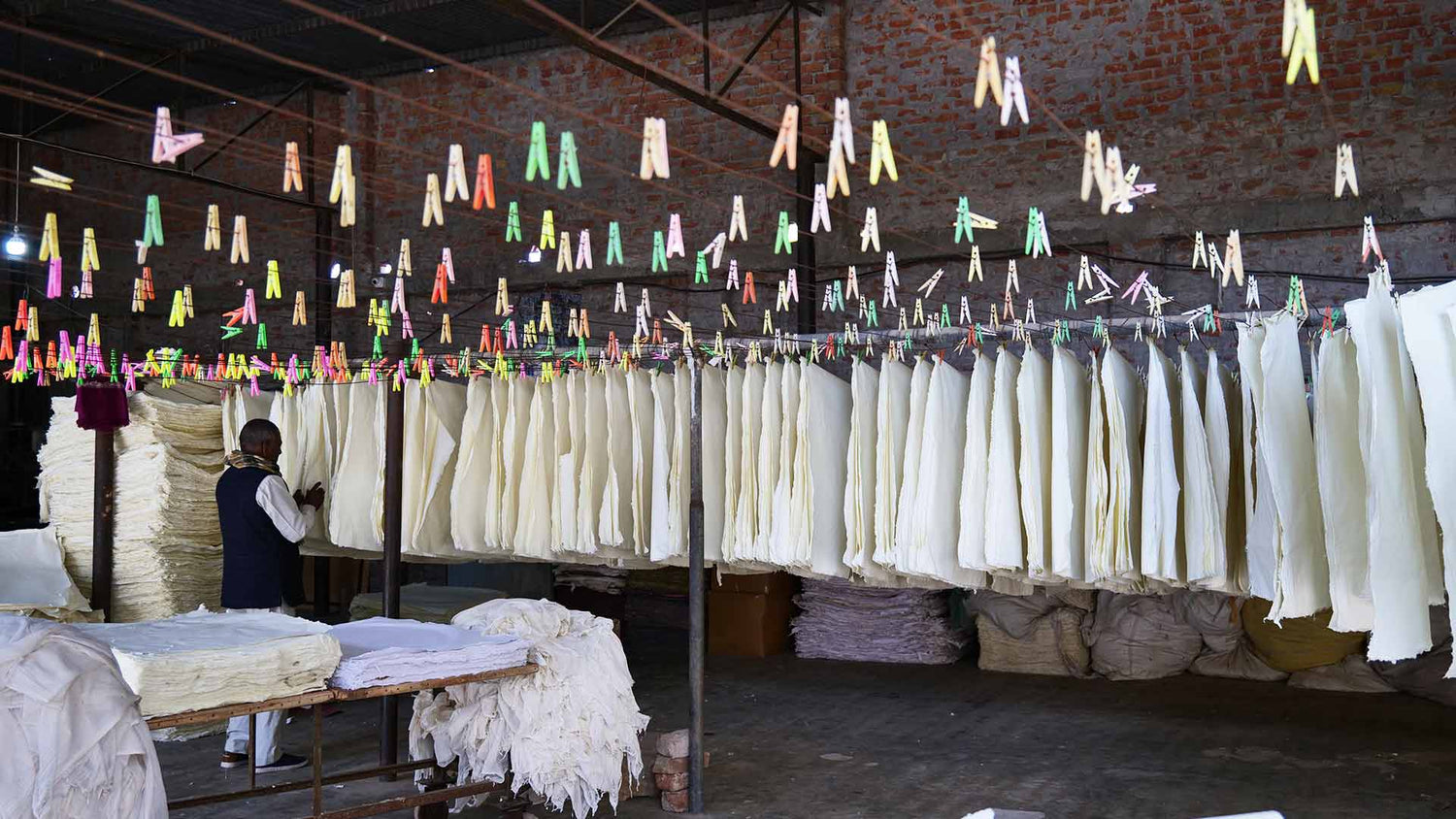 Many sheets of handmade paper held by multi coloured plastic pegs on clothes lines inside a factory.